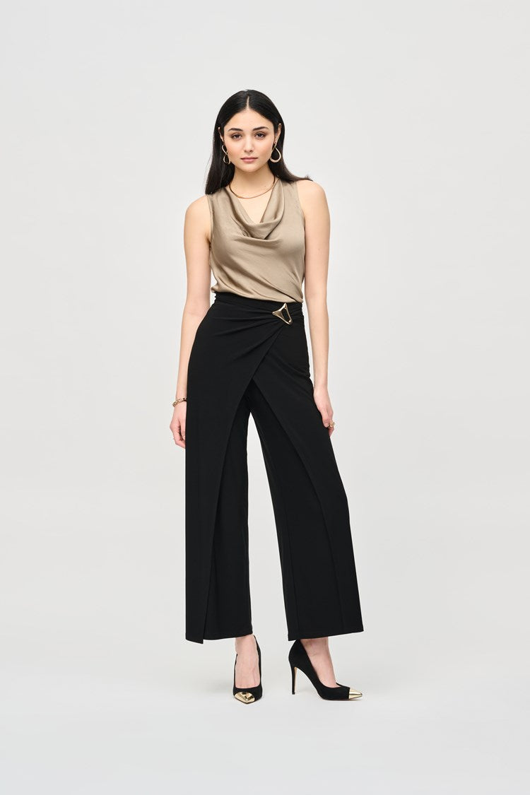 A woman stands confidently against a plain background. She is wearing a long-sleeved, geometric-patterned jacket over a black top and Joseph Ribkoff’s Silky Knit Wide-Leg Wrap Pants with slits. She completes her look with black pointed-toe heels, large hoop earrings, and an eye-catching asymmetrical wrap design.