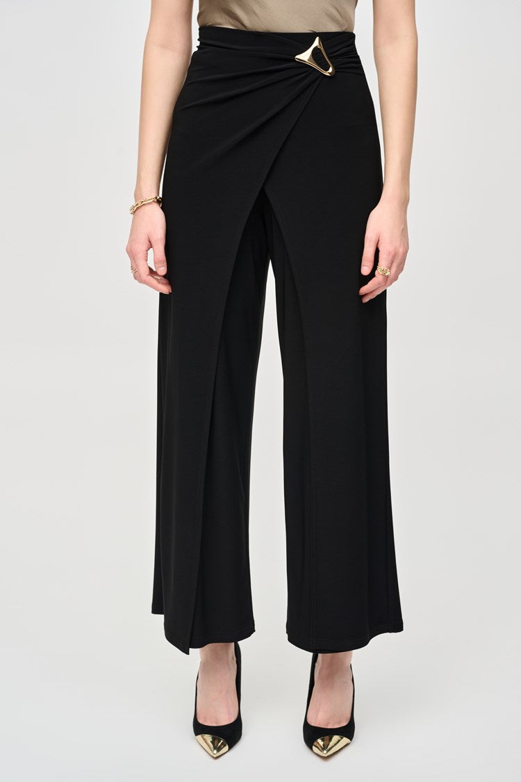 A woman stands confidently against a plain background. She is wearing a long-sleeved, geometric-patterned jacket over a black top and Joseph Ribkoff’s Silky Knit Wide-Leg Wrap Pants with slits. She completes her look with black pointed-toe heels, large hoop earrings, and an eye-catching asymmetrical wrap design.