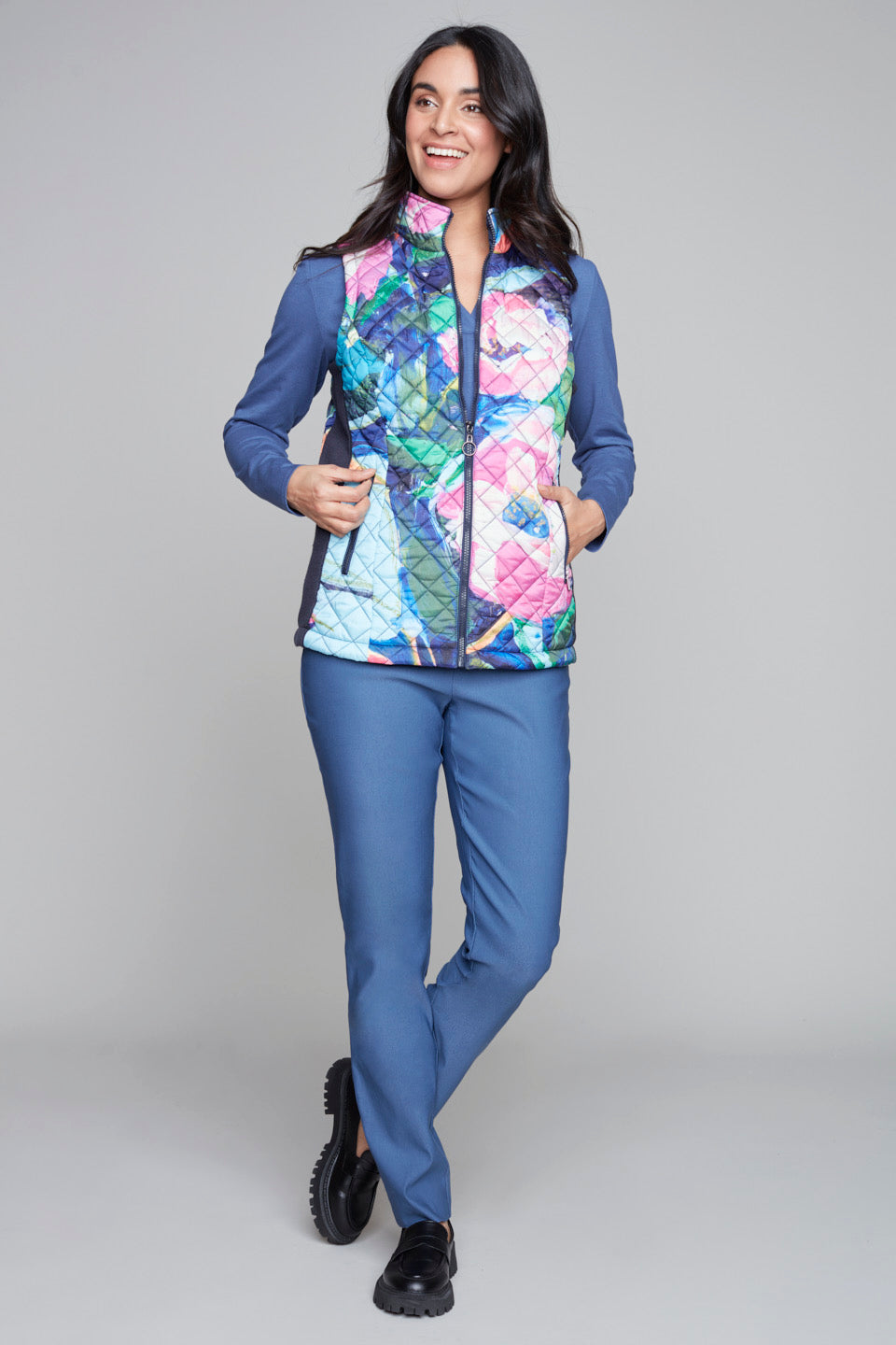 A woman with long dark hair stands against a gray background, donning the vibrant and stylish Woven Vest 91715 Clare Desjardins by Claire Desjardins over a blue long-sleeve top. One hand is in her pocket, and she looks confidently at the camera.