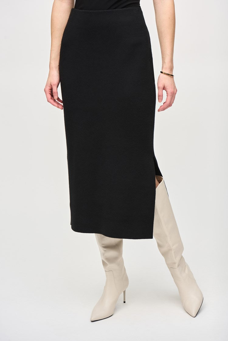 A person with long, red hair is standing against a plain white background. They are wearing a black, loose-fitting top with pocket details and the Joseph Ribkoff Sweater Knit Midi Skirt (style 243967) featuring a side slit and covered elastic waistband. They are also wearing white, heeled boots and dangling earrings.