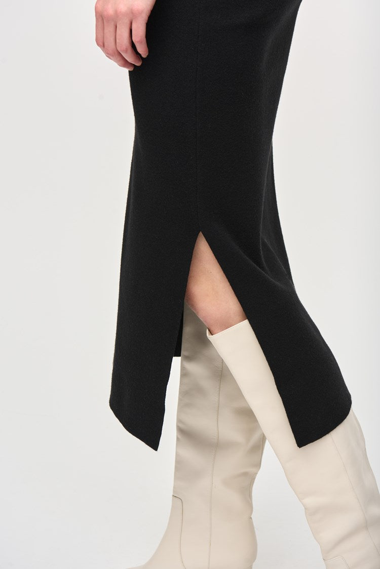 A person with long, red hair is standing against a plain white background. They are wearing a black, loose-fitting top with pocket details and the Joseph Ribkoff Sweater Knit Midi Skirt (style 243967) featuring a side slit and covered elastic waistband. They are also wearing white, heeled boots and dangling earrings.