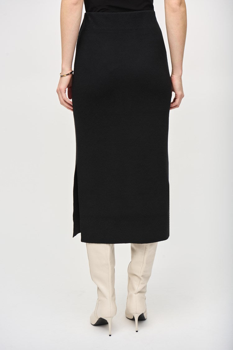 A person with long, red hair is standing against a plain white background. They are wearing a black, loose-fitting top with pocket details and the Joseph Ribkoff Sweater Knit Midi Skirt (style 243967) featuring a side slit and covered elastic waistband. They are also wearing white, heeled boots and dangling earrings.