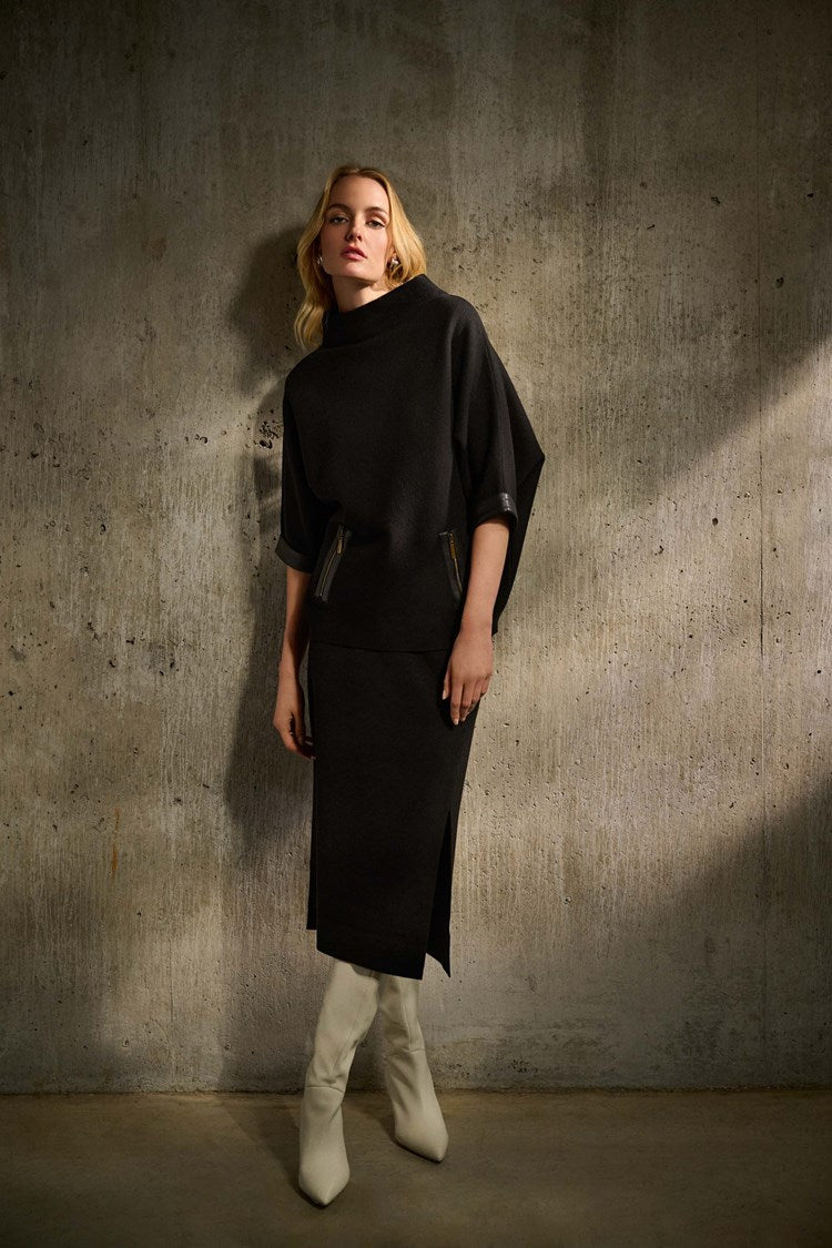 A person with long, red hair is standing against a plain white background. They are wearing a black, loose-fitting top with pocket details and the Joseph Ribkoff Sweater Knit Midi Skirt (style 243967) featuring a side slit and covered elastic waistband. They are also wearing white, heeled boots and dangling earrings.
