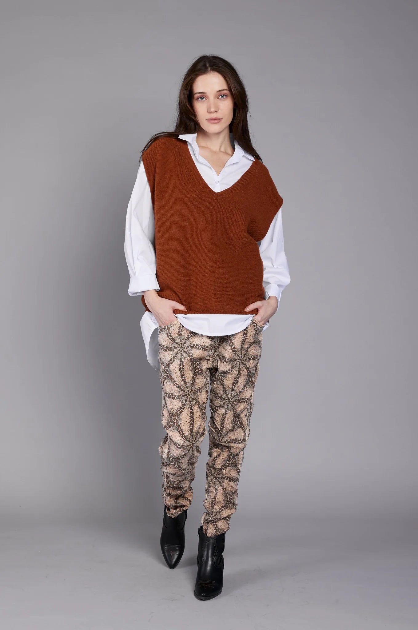 A woman with long dark hair stands against a plain gray background. She wears a white button-up shirt underneath the ELISSIA SL11316 Top, which is an oversized rust-colored sleeveless sweater, paired with patterned beige pants. Her hands are partially in her pockets, and she gazes confidently at the camera.