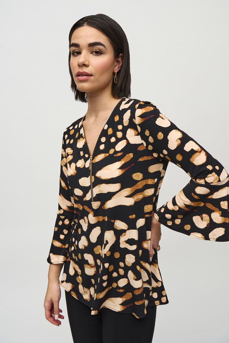 A woman with shoulder-length dark hair is standing against a plain backdrop. She is wearing the Silky Knit Animal Print Tunic by Joseph Ribkoff, paired with black high-waisted trousers that have gold buttons on the front and black heeled boots.