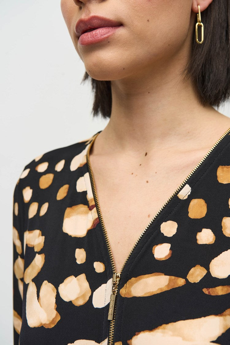 A woman with shoulder-length dark hair is standing against a plain backdrop. She is wearing the Silky Knit Animal Print Tunic by Joseph Ribkoff, paired with black high-waisted trousers that have gold buttons on the front and black heeled boots.
