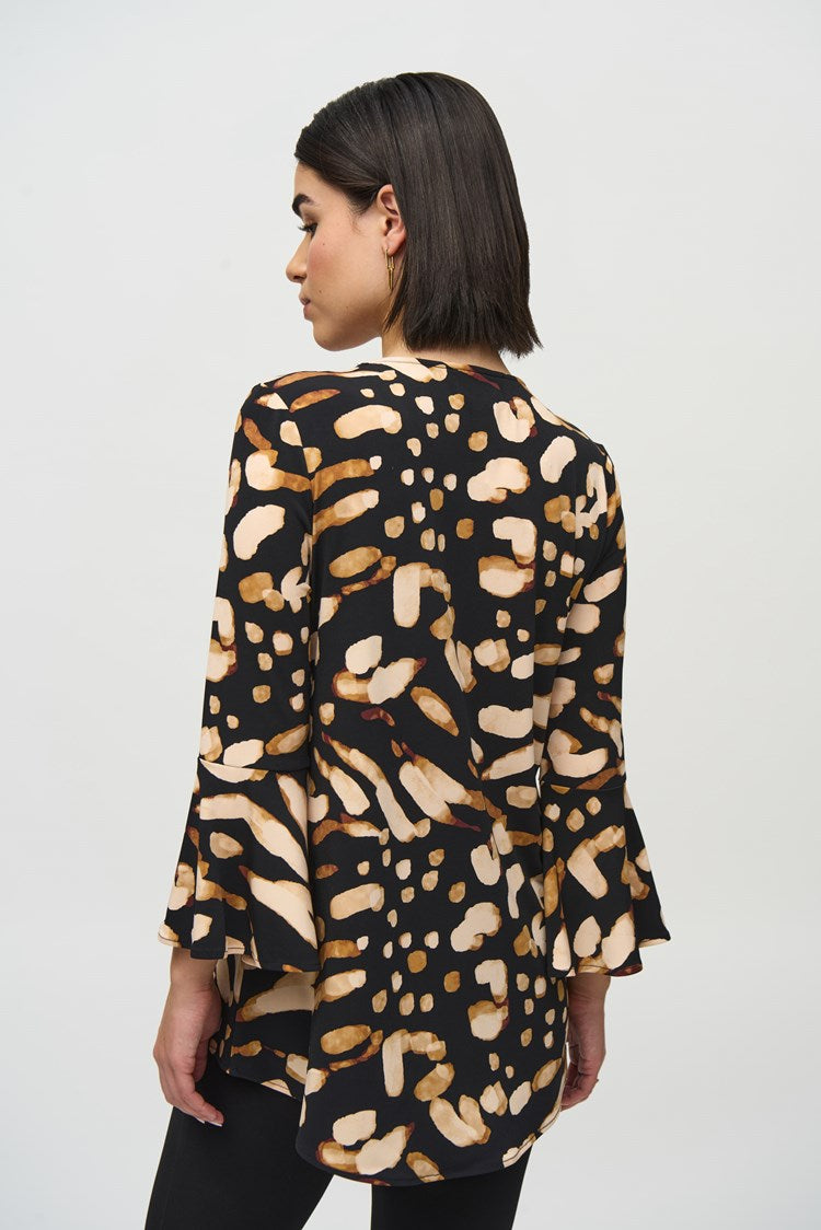 A woman with shoulder-length dark hair is standing against a plain backdrop. She is wearing the Silky Knit Animal Print Tunic by Joseph Ribkoff, paired with black high-waisted trousers that have gold buttons on the front and black heeled boots.