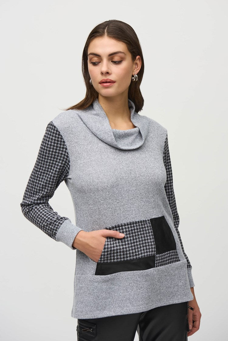 A person with long hair stands against a plain background wearing a stylish ensemble. The outfit includes the Joseph Ribkoff Sweater Knit Houndstooth Tunic, featuring a gray cowl-neck design with black and white houndstooth print details on the sleeves and pocket, paired with black pants and ankle boots. They touch their head with one hand.