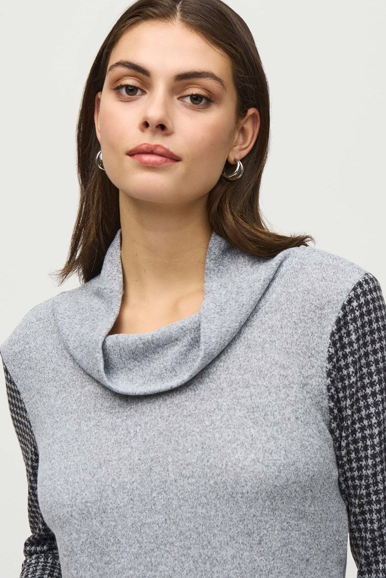 A person with long hair stands against a plain background wearing a stylish ensemble. The outfit includes the Joseph Ribkoff Sweater Knit Houndstooth Tunic, featuring a gray cowl-neck design with black and white houndstooth print details on the sleeves and pocket, paired with black pants and ankle boots. They touch their head with one hand.