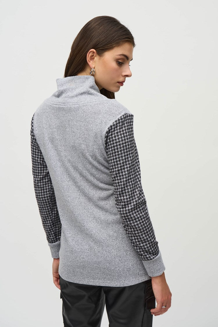 A person with long hair stands against a plain background wearing a stylish ensemble. The outfit includes the Joseph Ribkoff Sweater Knit Houndstooth Tunic, featuring a gray cowl-neck design with black and white houndstooth print details on the sleeves and pocket, paired with black pants and ankle boots. They touch their head with one hand.