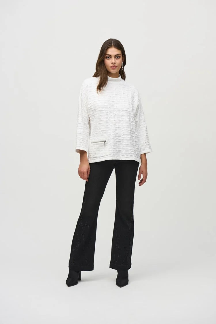 A woman with long, brown hair wears a Jacquard Sweater Knit Boxy Top by Joseph Ribkoff, featuring three-quarter dolman sleeves and a zippered pocket on one side. She stands against a plain light gray background, gazing downward with a neutral expression.