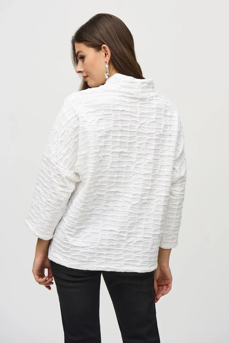 A woman with long, brown hair wears a Jacquard Sweater Knit Boxy Top by Joseph Ribkoff, featuring three-quarter dolman sleeves and a zippered pocket on one side. She stands against a plain light gray background, gazing downward with a neutral expression.