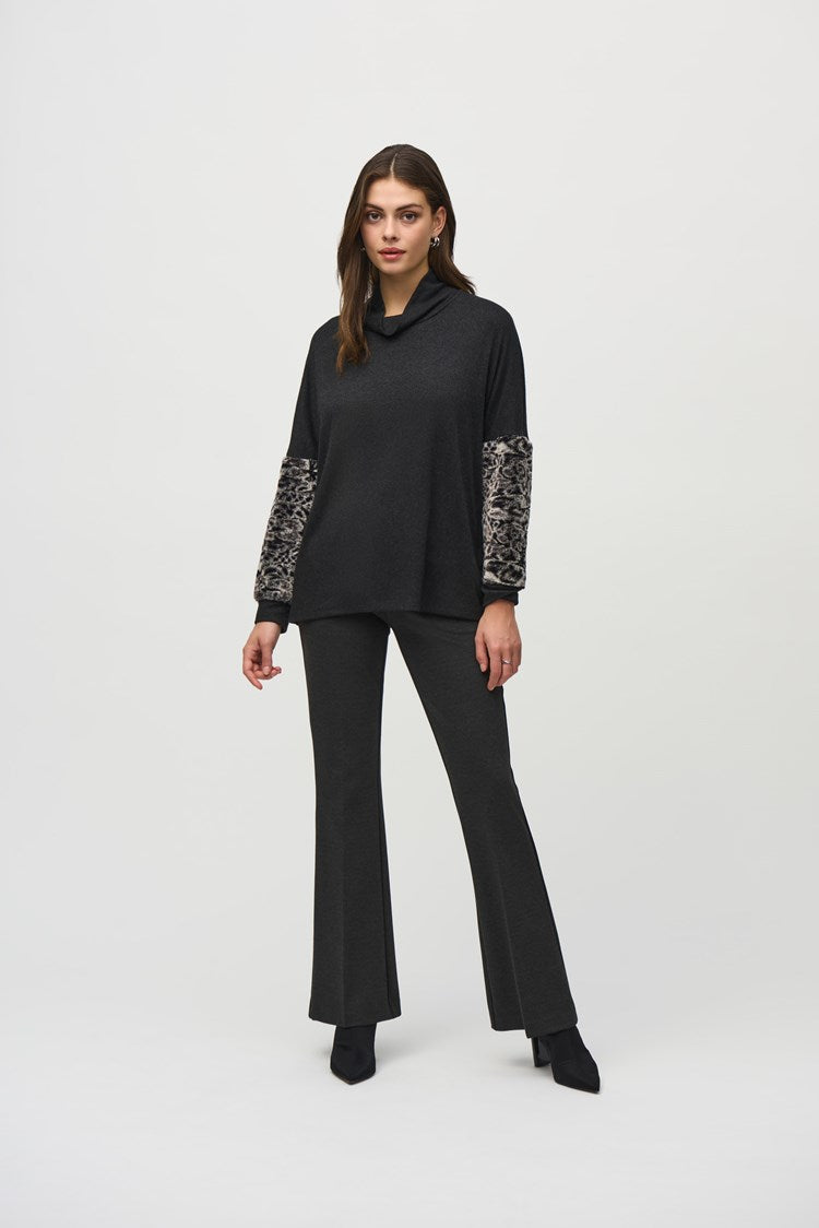 A woman stands against a plain background, wearing a black Joseph Ribkoff Sweater Knit Top With Faux Fur Sleeves (Product Code: 244247) and dark grey pants. She has long, dark hair and wears black pointed-toe shoes. Her left hand is in her pocket as she looks at the camera with a neutral expression.