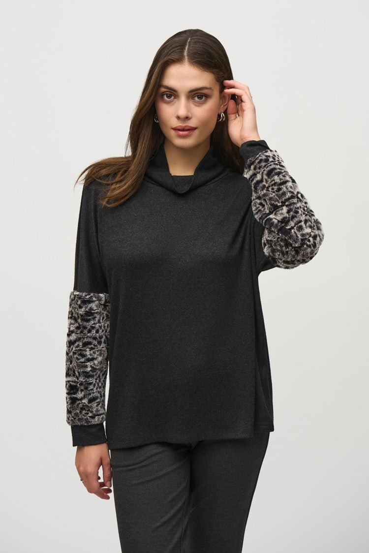 A woman stands against a plain background, wearing a black Joseph Ribkoff Sweater Knit Top With Faux Fur Sleeves (Product Code: 244247) and dark grey pants. She has long, dark hair and wears black pointed-toe shoes. Her left hand is in her pocket as she looks at the camera with a neutral expression.