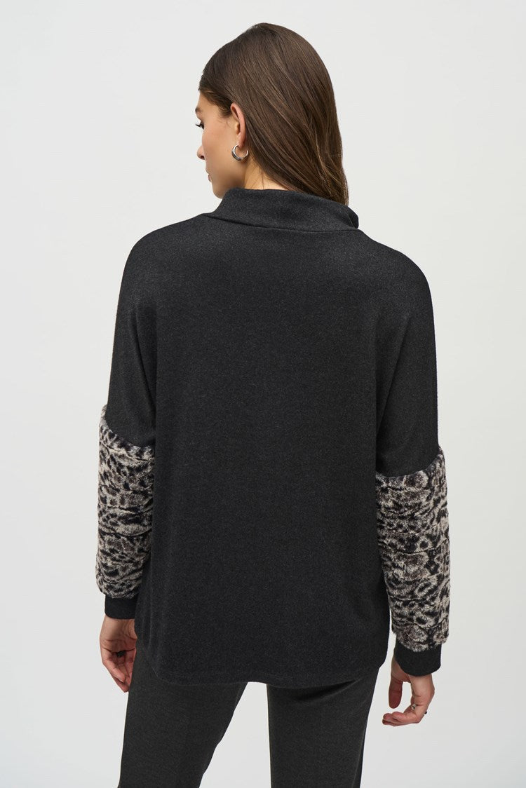 A woman stands against a plain background, wearing a black Joseph Ribkoff Sweater Knit Top With Faux Fur Sleeves (Product Code: 244247) and dark grey pants. She has long, dark hair and wears black pointed-toe shoes. Her left hand is in her pocket as she looks at the camera with a neutral expression.
