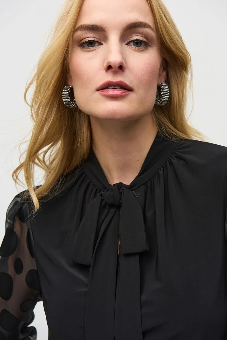 A woman stands confidently against a white background. She is wearing the Joseph Ribkoff Silky Knit Top with Dot Burnout Sleeves 244019, featuring sheer sleeves with dot burnout detailing and a bow tie at the neck. She pairs it with black wide-leg jeans and silver high-heeled shoes. Her light-blonde hair is styled in loose waves.