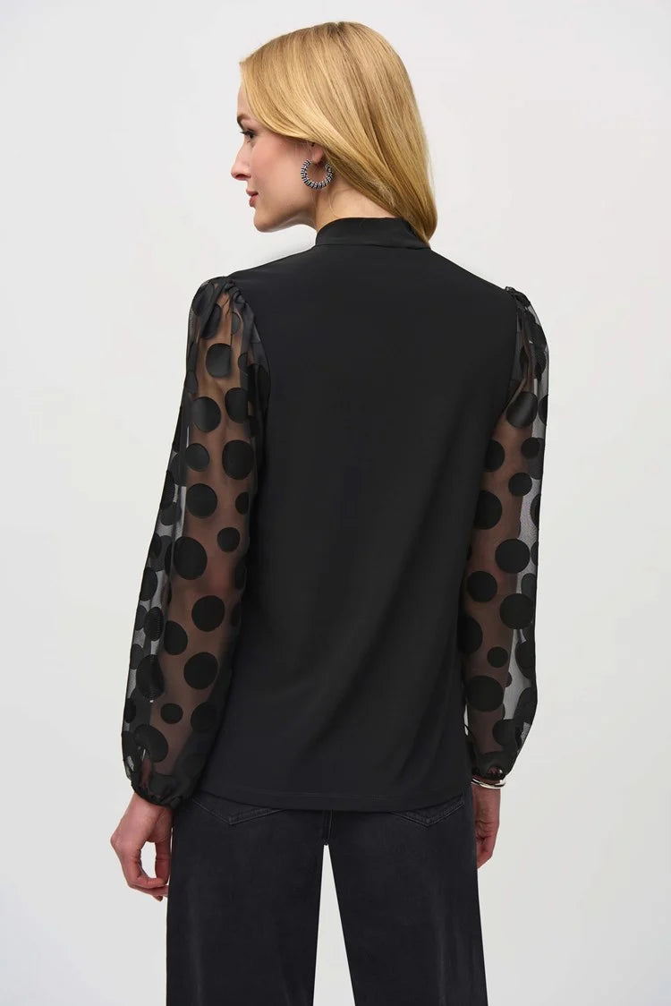 A woman stands confidently against a white background. She is wearing the Joseph Ribkoff Silky Knit Top with Dot Burnout Sleeves 244019, featuring sheer sleeves with dot burnout detailing and a bow tie at the neck. She pairs it with black wide-leg jeans and silver high-heeled shoes. Her light-blonde hair is styled in loose waves.