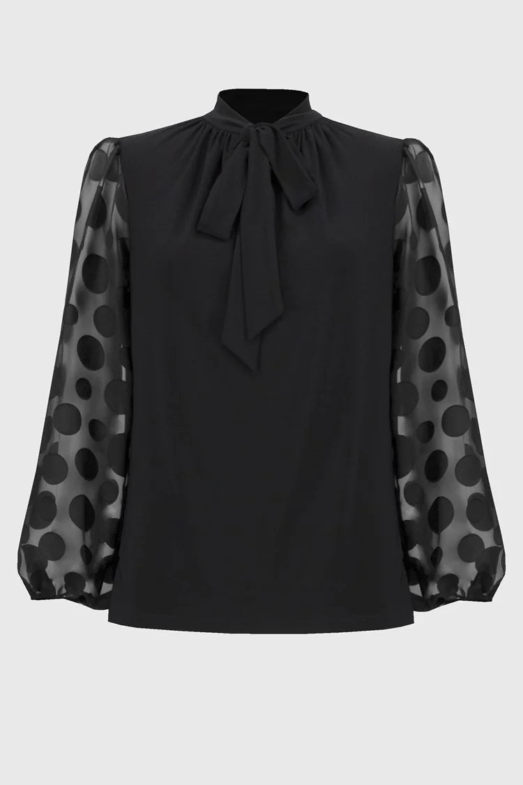 A woman stands confidently against a white background. She is wearing the Joseph Ribkoff Silky Knit Top with Dot Burnout Sleeves 244019, featuring sheer sleeves with dot burnout detailing and a bow tie at the neck. She pairs it with black wide-leg jeans and silver high-heeled shoes. Her light-blonde hair is styled in loose waves.