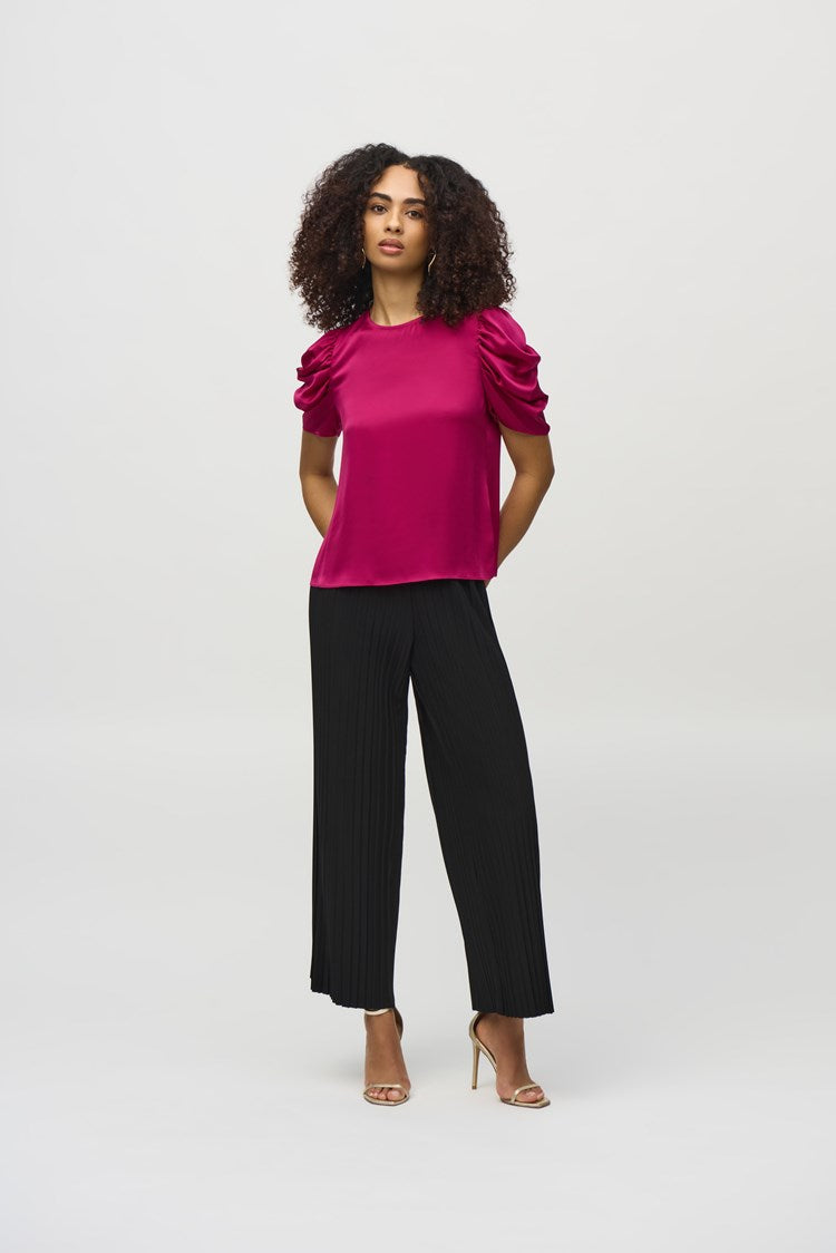 A woman with curly hair, wearing the Joseph Ribkoff Satin Straight Top With Shirred Sleeves (style number 244023) and black pleated pants, stands against a plain white background. She gazes directly at the camera with a neutral expression.