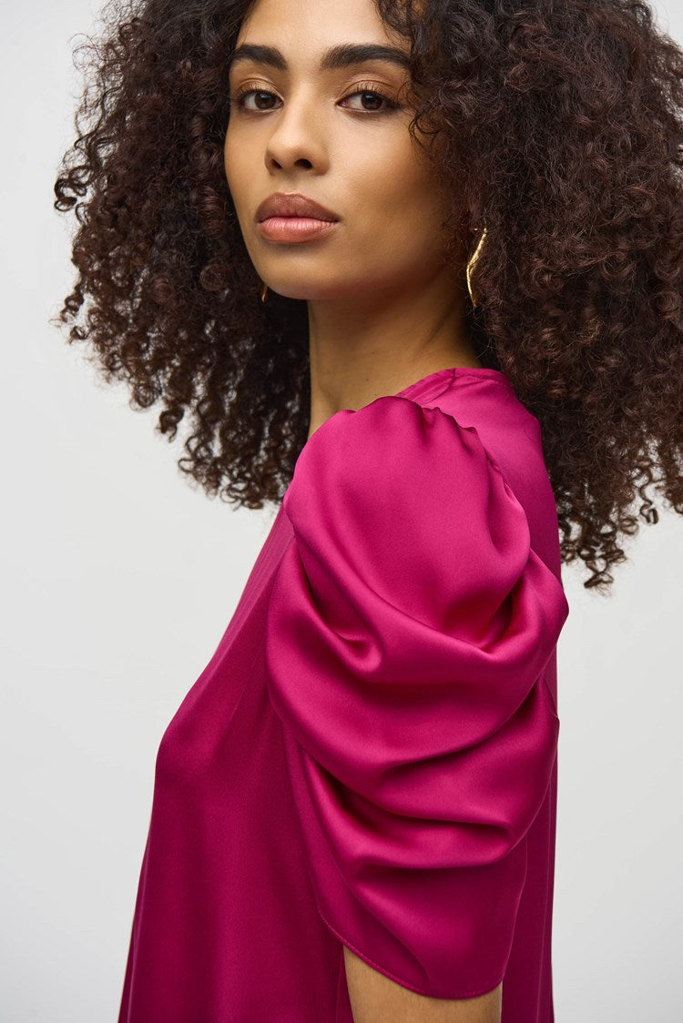 A woman with curly hair, wearing the Joseph Ribkoff Satin Straight Top With Shirred Sleeves (style number 244023) and black pleated pants, stands against a plain white background. She gazes directly at the camera with a neutral expression.