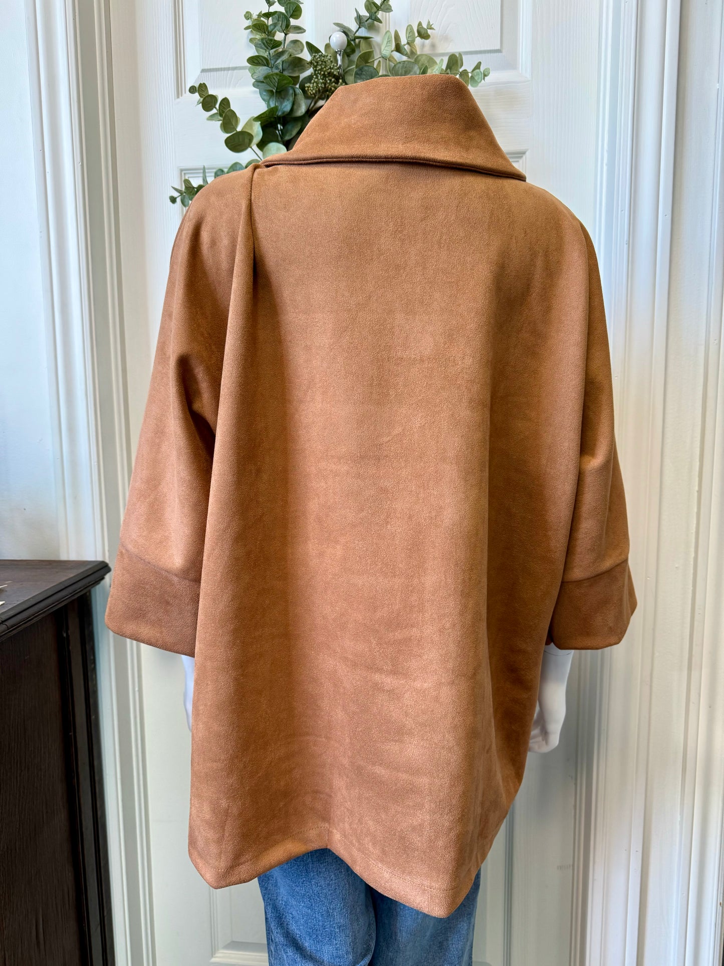 A mannequin showcases the ELISSIA BC12762 Shawl 12762 - Elissia, a camel-colored, suede-like jacket featuring wide sleeves and a distinctive buttoned collar. This luxurious jacket, complete with two front pockets, is elegantly paired with blue jeans. In the background, an assortment of clothing items and some greenery are hanging on a door.