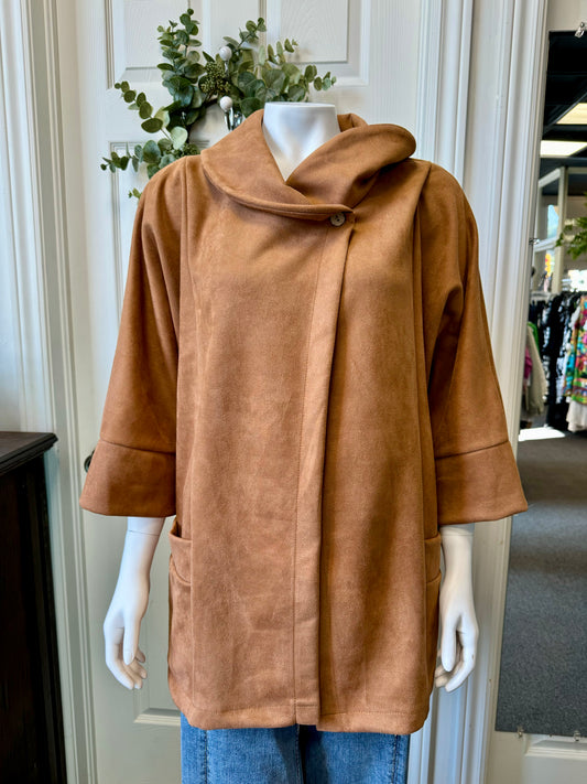 A mannequin showcases the ELISSIA BC12762 Shawl 12762 - Elissia, a camel-colored, suede-like jacket featuring wide sleeves and a distinctive buttoned collar. This luxurious jacket, complete with two front pockets, is elegantly paired with blue jeans. In the background, an assortment of clothing items and some greenery are hanging on a door.