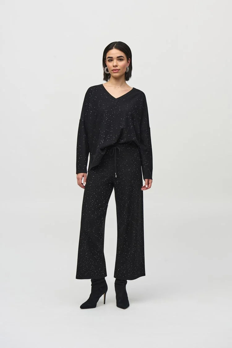 A person with shoulder-length dark hair is wearing the Joseph Ribkoff Sequined Sweater Knit Boxy Top-244921, paired with matching black pants featuring a white speckled pattern. Their hands are clasped together, and they are looking slightly to the side while standing against a plain, light-colored background.