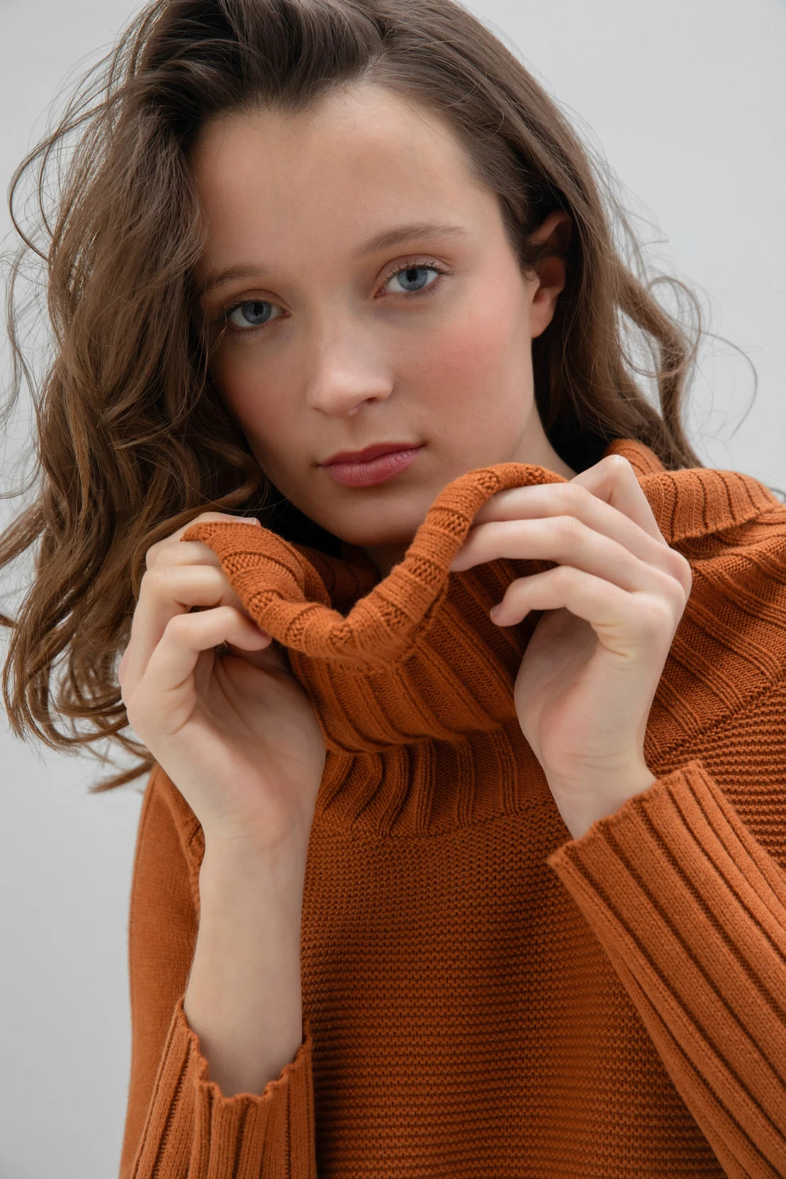 A woman with wavy hair is standing in a minimalistic setting, wearing the Marble Sweater-7604 by MARBLE, a caramel-colored sweater with a draped collar, paired with a colorful, patterned skirt. She is also wearing black ankle boots. The background is plain and light gray.