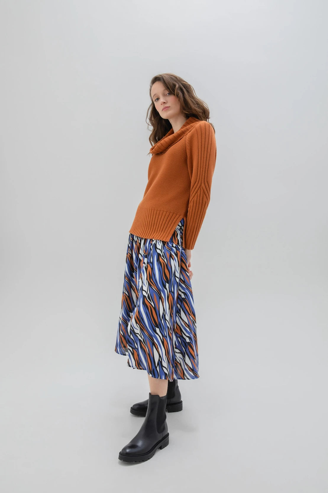 A woman with wavy hair is standing in a minimalistic setting, wearing the Marble Sweater-7604 by MARBLE, a caramel-colored sweater with a draped collar, paired with a colorful, patterned skirt. She is also wearing black ankle boots. The background is plain and light gray.