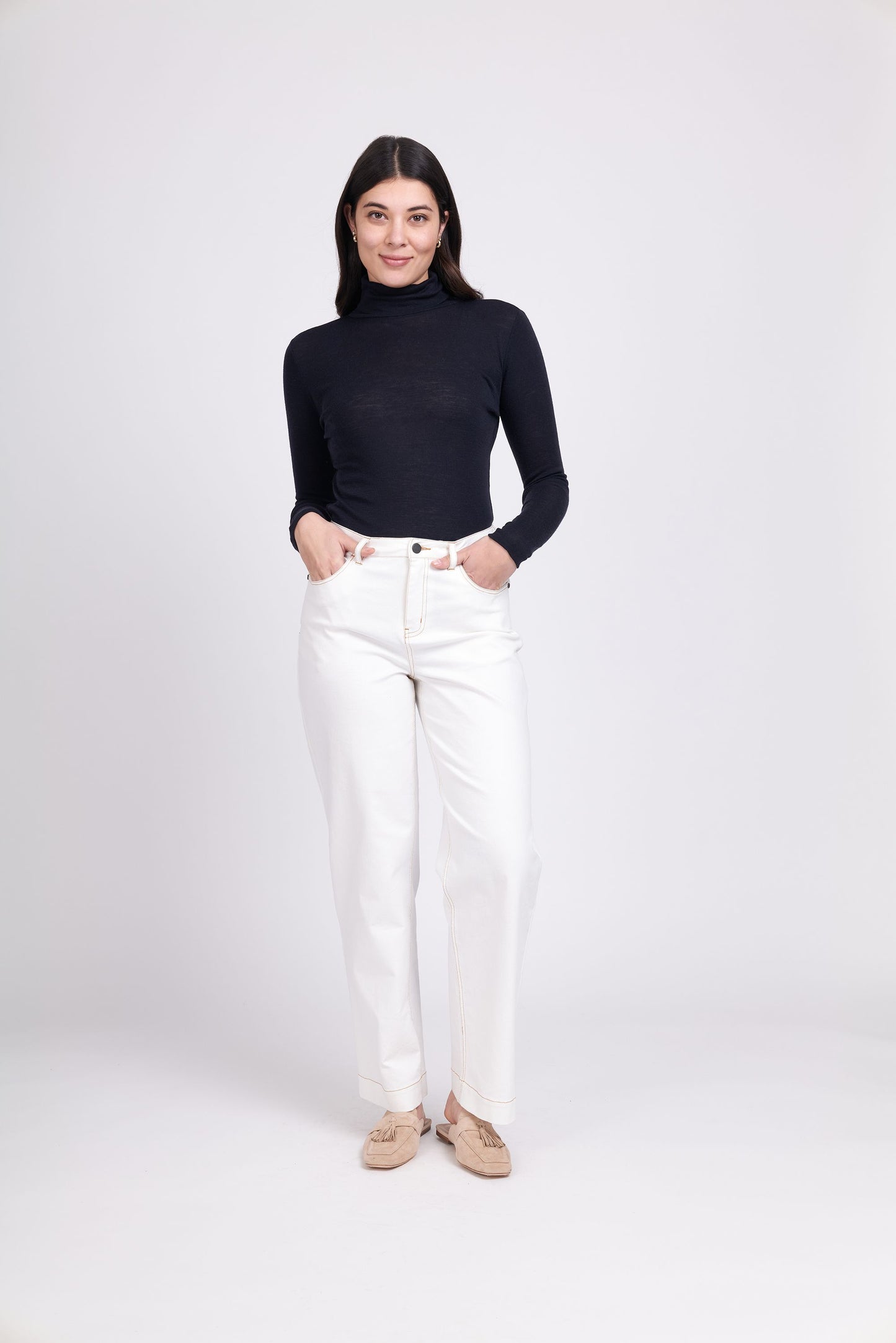 A person with long dark hair stands against a plain light background, wearing a FOIL Fine Merino Polo Neck Top-Navy paired with high-waisted white pants. Their hands are in their pockets, and they are wearing beige shoes. They are smiling gently while facing the camera.