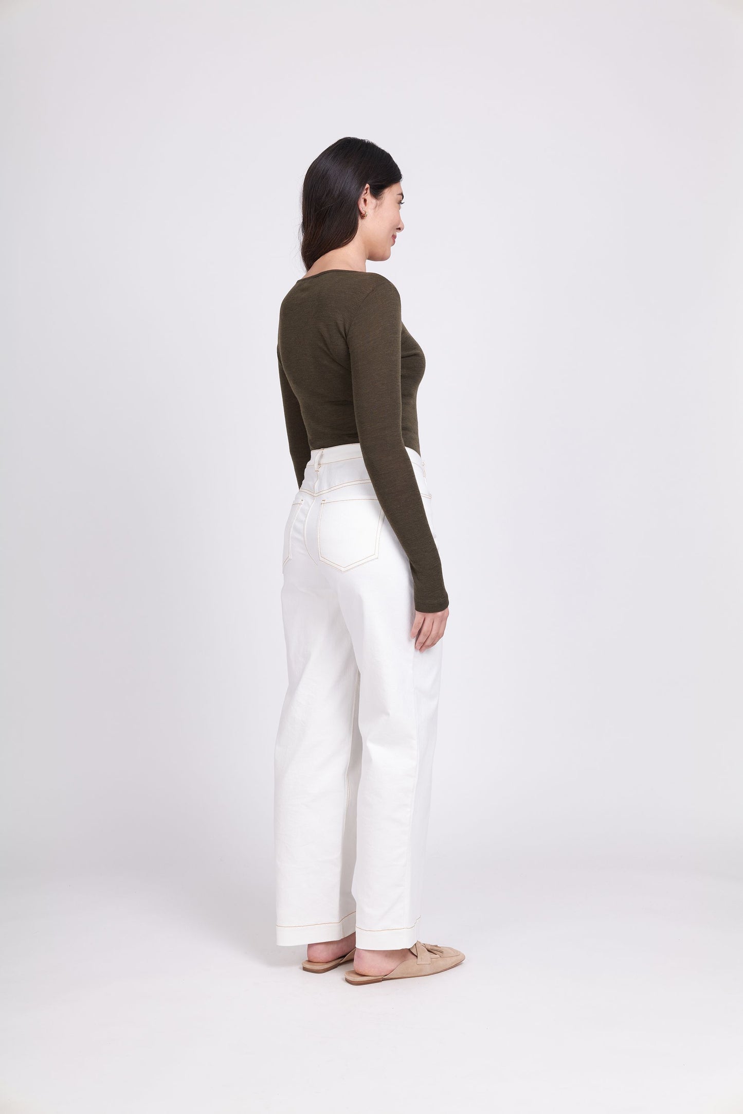 A woman stands against a white background wearing the FOIL Fine Merino Crew Neck Top in khaki marl green and high-waisted white jeans. She has long dark hair and is smiling slightly, with her hands by her sides. She is also wearing beige flats.