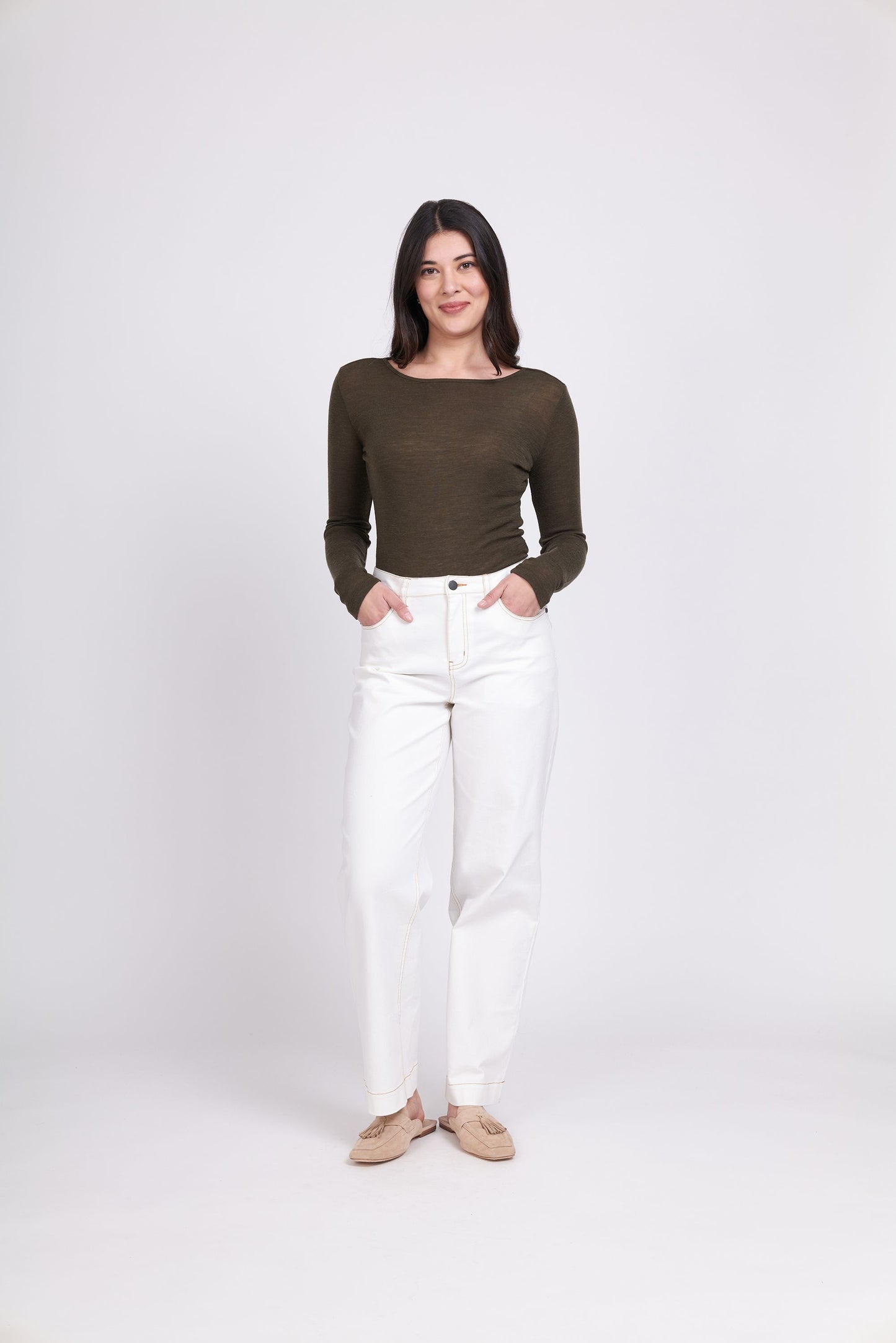 A woman stands against a white background wearing the FOIL Fine Merino Crew Neck Top in khaki marl green and high-waisted white jeans. She has long dark hair and is smiling slightly, with her hands by her sides. She is also wearing beige flats.