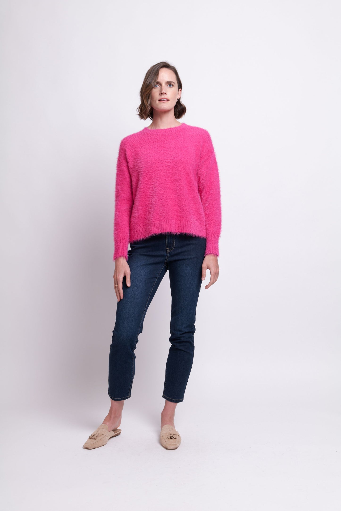A woman stands against a plain background wearing the bright pink "Fluff Love Sweater" from FOIL, paired with dark blue jeans and beige shoes. With short brown hair and a subtle sideways gaze, she embodies the perfect addition to a cozy winter wardrobe.