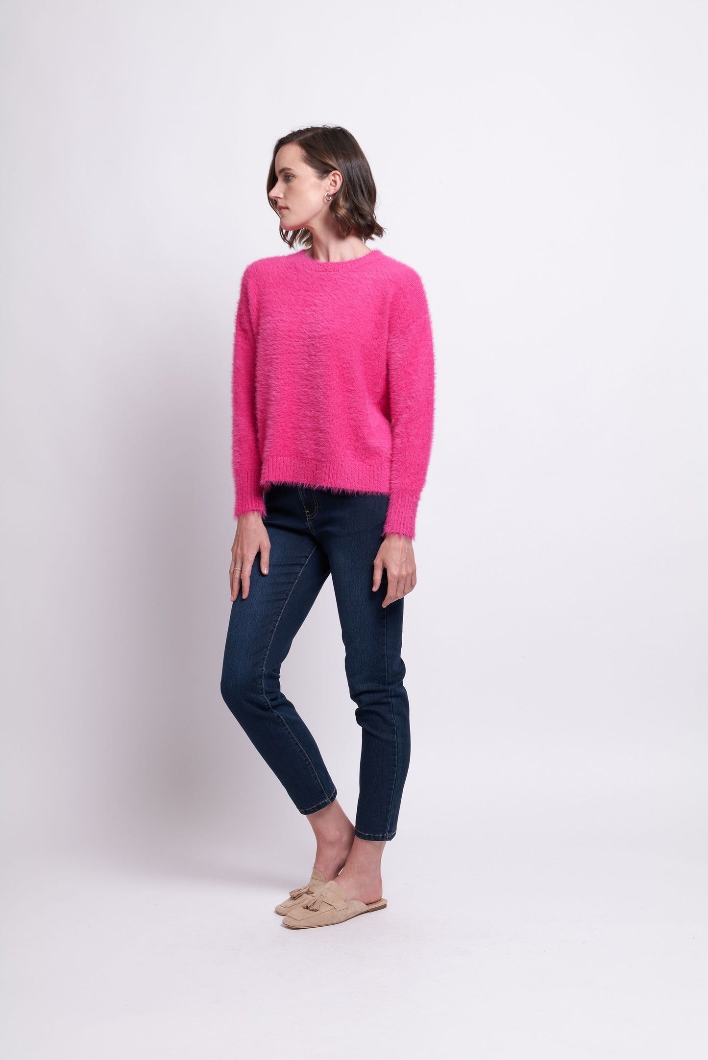 A woman stands against a plain background wearing the bright pink "Fluff Love Sweater" from FOIL, paired with dark blue jeans and beige shoes. With short brown hair and a subtle sideways gaze, she embodies the perfect addition to a cozy winter wardrobe.