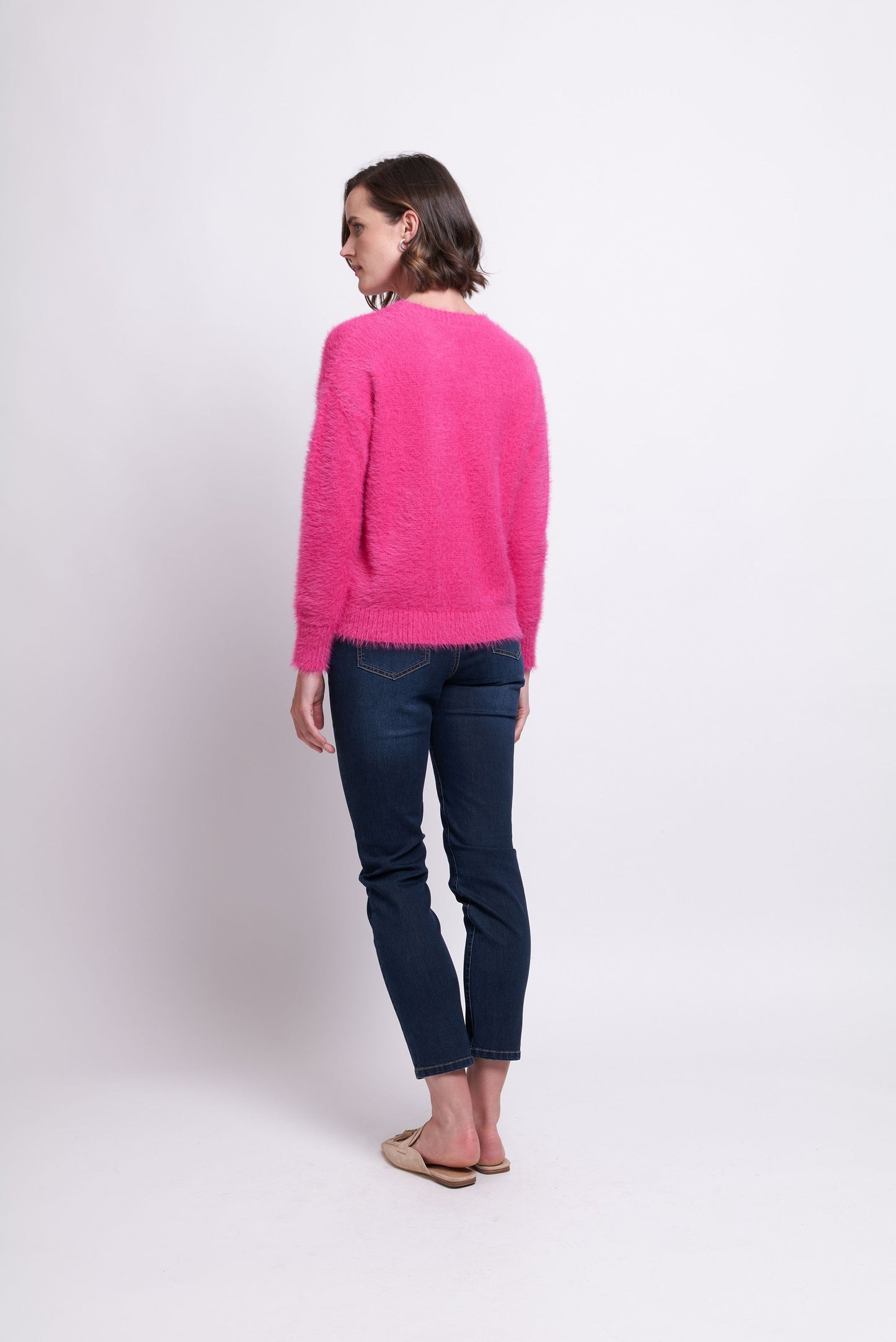 A woman stands against a plain background wearing the bright pink "Fluff Love Sweater" from FOIL, paired with dark blue jeans and beige shoes. With short brown hair and a subtle sideways gaze, she embodies the perfect addition to a cozy winter wardrobe.