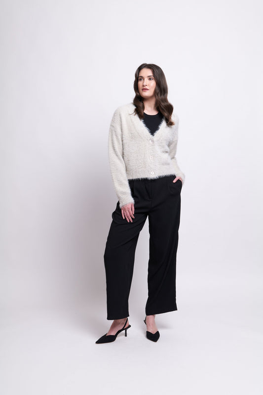 Against a plain white background, a woman is seen wearing the stylish and comfortable FOIL Good Fluff Cardigan—an elegant, fluffy beige piece layered over a black top. She has paired it with black wide-leg trousers and black pointed-toe heels. With her hands casually tucked into her pockets, she showcases her long, wavy dark hair effortlessly.