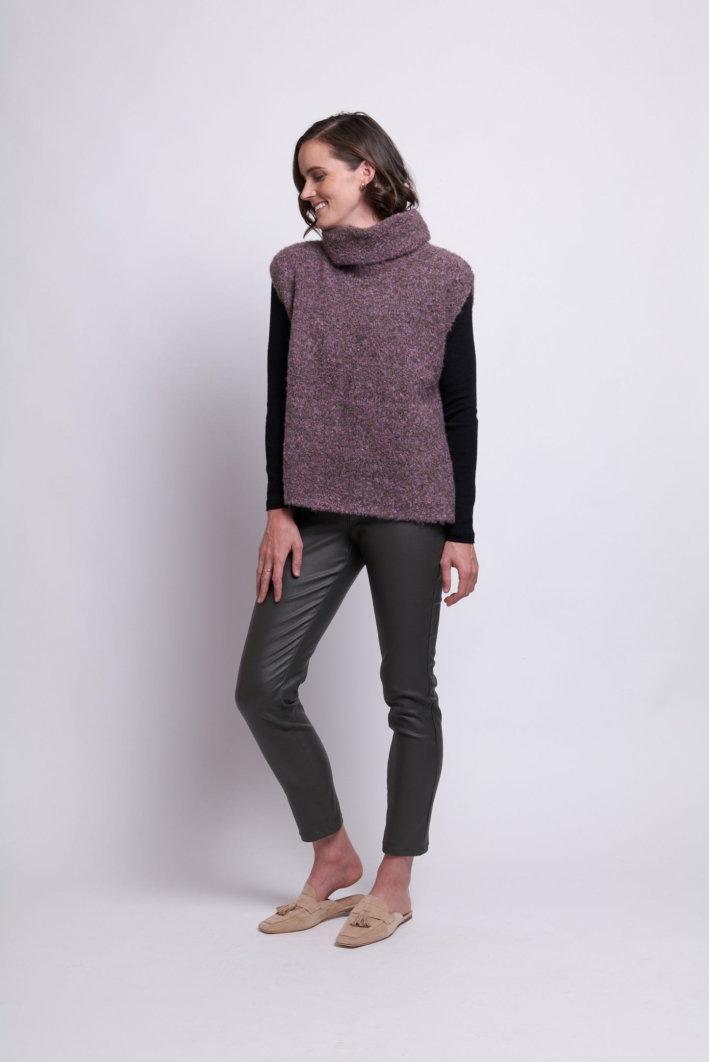 A woman stands against a plain backdrop, showcasing the "Scrunch Time Pop Over" by FOIL. She pairs the sleeveless, purple turtleneck sweater with a black long-sleeve shirt and completes her outfit with dark gray slim-fit pants and stylish beige loafers. Her short brown hair frames her face as she gazes forward with her hands clasped in front. The versatile color palette exudes effortless elegance.