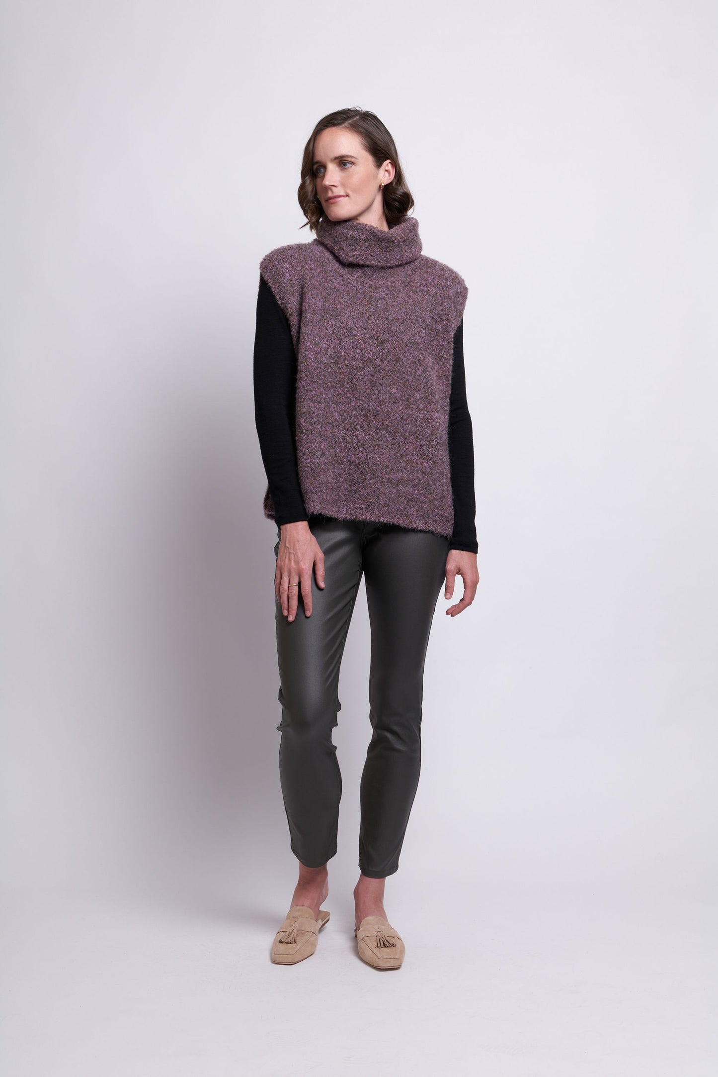 A woman stands against a plain backdrop, showcasing the "Scrunch Time Pop Over" by FOIL. She pairs the sleeveless, purple turtleneck sweater with a black long-sleeve shirt and completes her outfit with dark gray slim-fit pants and stylish beige loafers. Her short brown hair frames her face as she gazes forward with her hands clasped in front. The versatile color palette exudes effortless elegance.