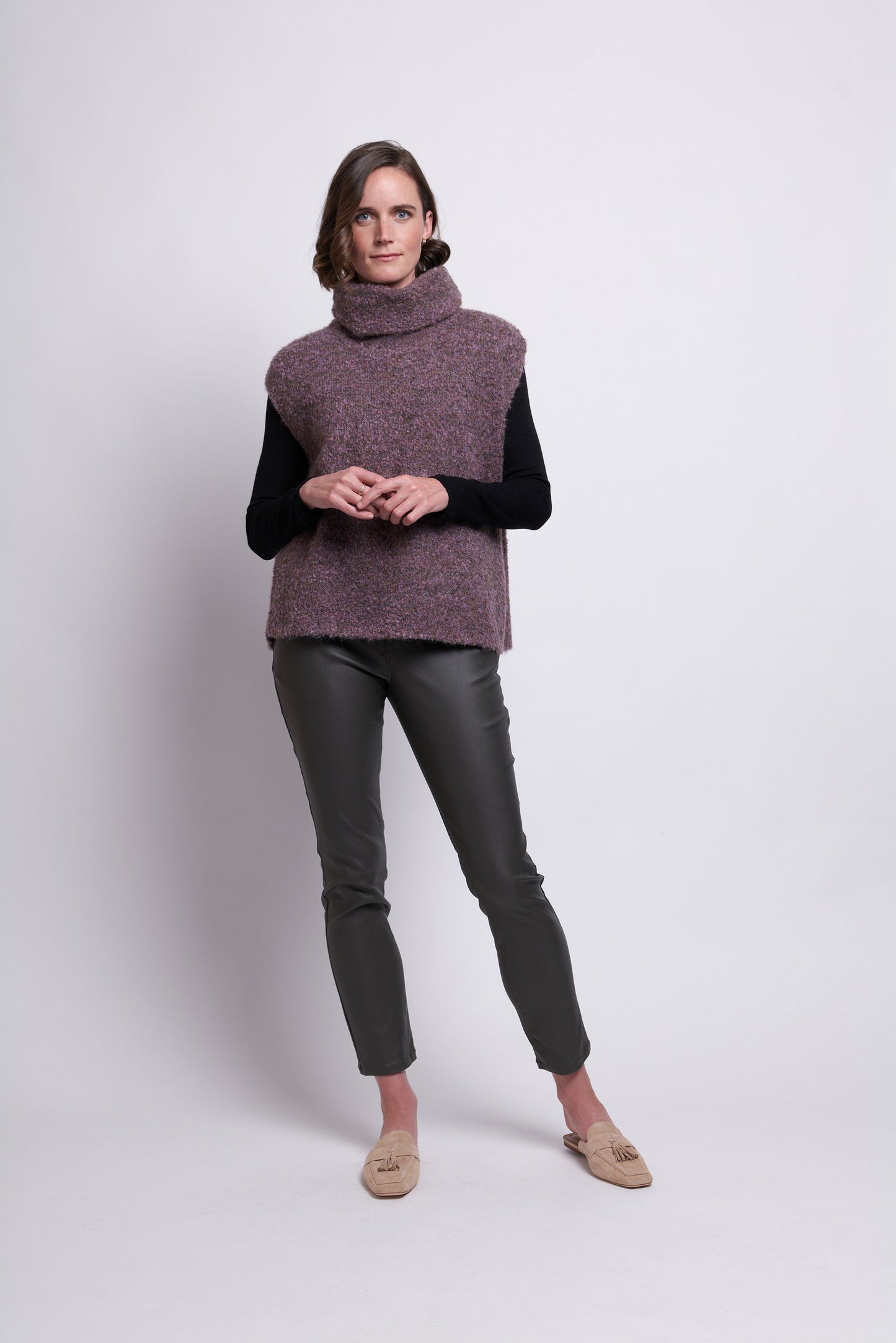 A woman stands against a plain white background, wearing the Scrunch Time Pop Over by FOIL, a chunky sleeveless turtleneck sweater, layered over a long-sleeve black shirt. She pairs this cozy ensemble with greenish-gray slim-fit pants and beige loafers, posing with her hands lightly clasped in front of her and exuding a stylish and comfortable vibe.