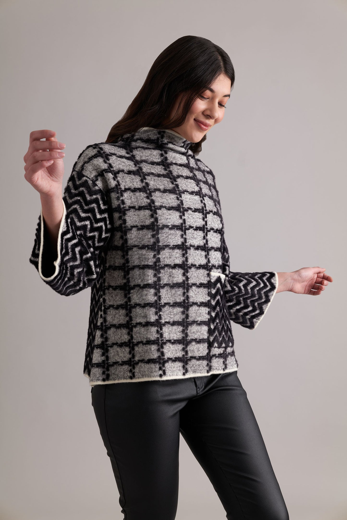 A woman with long dark hair, wearing a black and white FOIL Game Of Thrones Sweater with zigzag sleeves and black pants. She is looking down and smiling, with her arms slightly raised. The background is plain and light gray.
