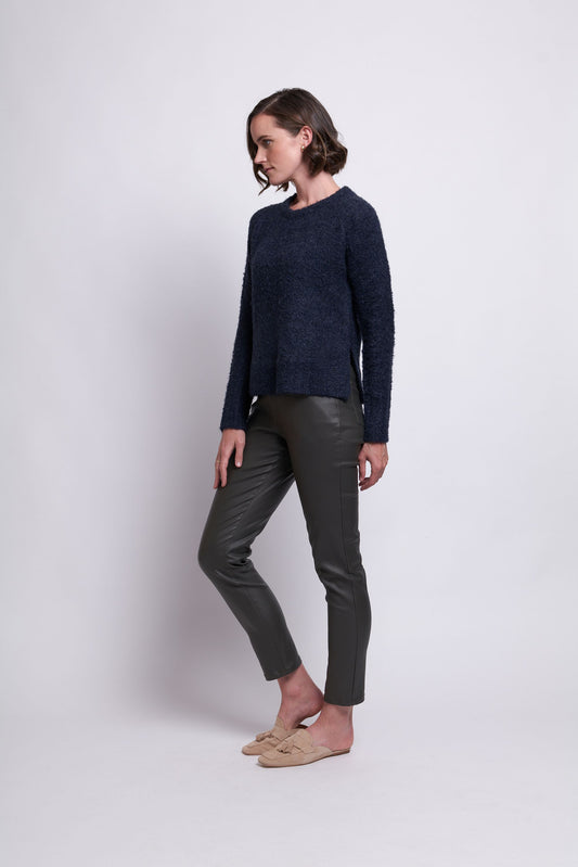 A person with short, dark hair stands against a plain white background. They are wearing a navy blue long-sleeve Raglan To Riches Sweater - FOIL made from recycled polyester, dark gray fitted pants, and beige slip-on shoes. They are looking down and slightly to their left with their arms relaxed by their sides.
