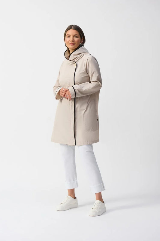 Wearing the Memory Hooded Trapeze Coat 251184 by Joseph Ribkoff, a person stands against a white background with relaxed arms. The light beige coat features contrast binding and a high collar, paired with white pants and sneakers for an effortlessly chic, vintage-inspired look.