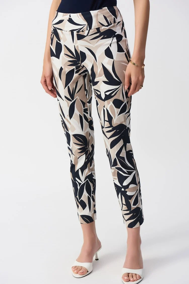 Wearing Joseph Ribkoff’s Millennium Tropical Print Slim Fit Pants 251001, which feature a high-waisted, bold black, white, and beige leaf pattern paired with a navy top and white open-toe heels. The outfit exudes elegance with one hand resting casually by their side.
