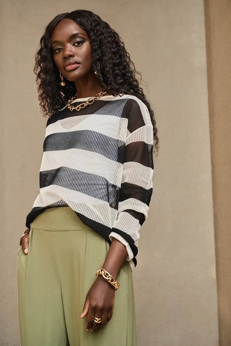 A person with long curly hair confidently poses in a Joseph Ribkoff Striped Sweater Knit Pullover 251922, paired with olive green pants. They accessorize with gold jewelry, including a chunky chain necklace and bracelets, against a neutral background.