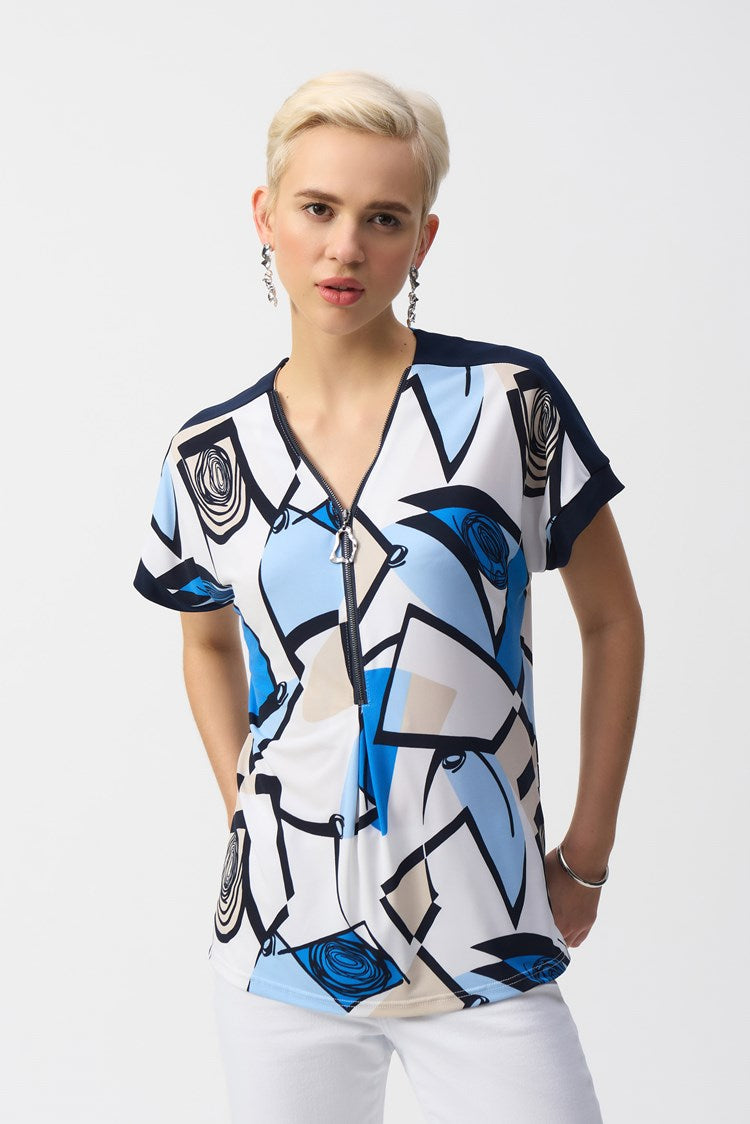 A short-haired person with blonde hair models the Joseph Ribkoff Silky Knit Geometric Print Straight Top 251246, featuring blue and white patterns and a front zipper, paired with white pants. They stand against a plain background, showcasing a neutral expression.