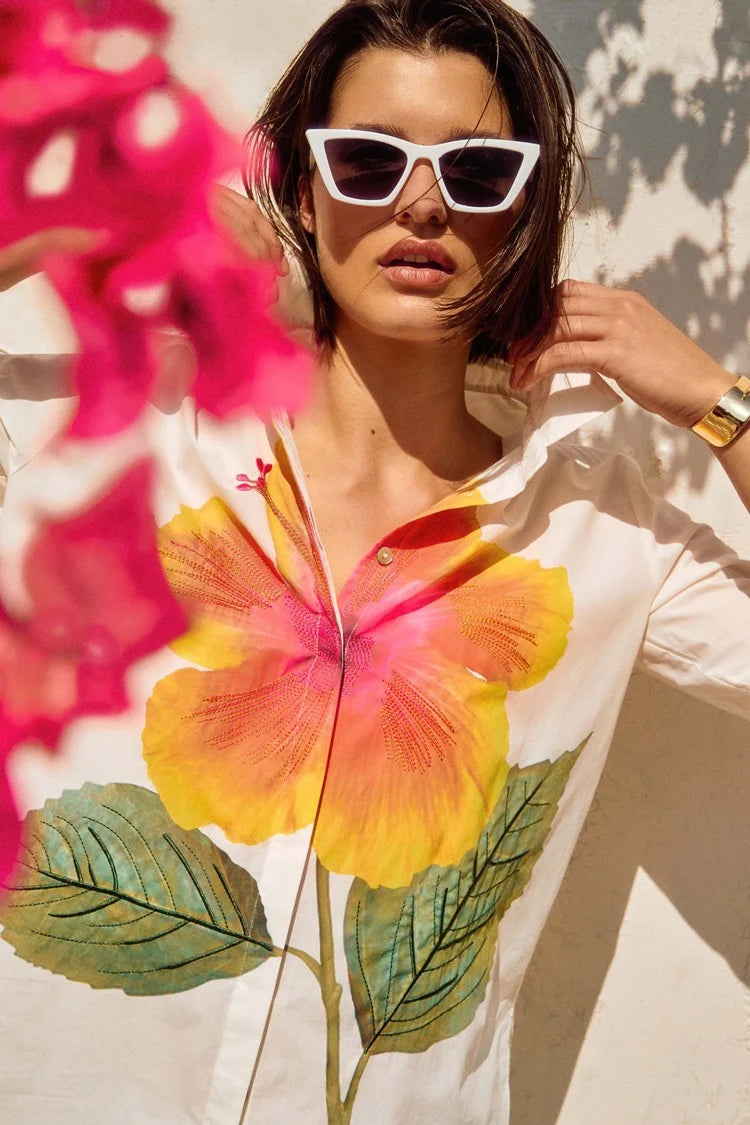 Someone in white sunglasses adjusts their collar, wearing a Joseph Ribkoff embroidered flower cotton shirt. Bright pink flowers blur in the foreground, adding vibrant touches to the sunny scene.