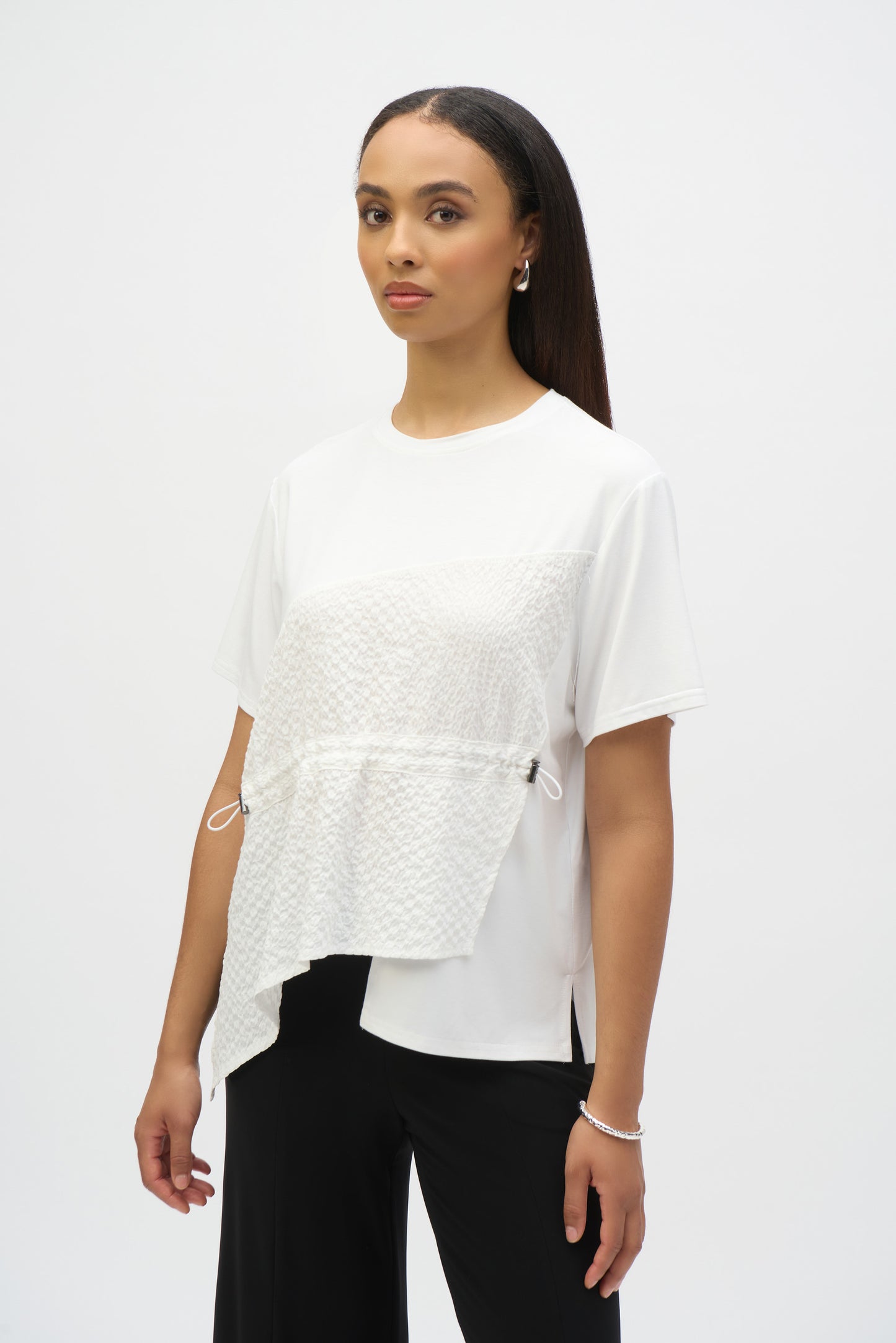 A woman effortlessly embodies casual chic, wearing Joseph Ribkoff's Jersey And Seersucker Asymmetrical Top 252117 against a plain white background, paired with black pants. Her long, dark hair complements hoop earrings and a bracelet, with her expression remaining neutral.
