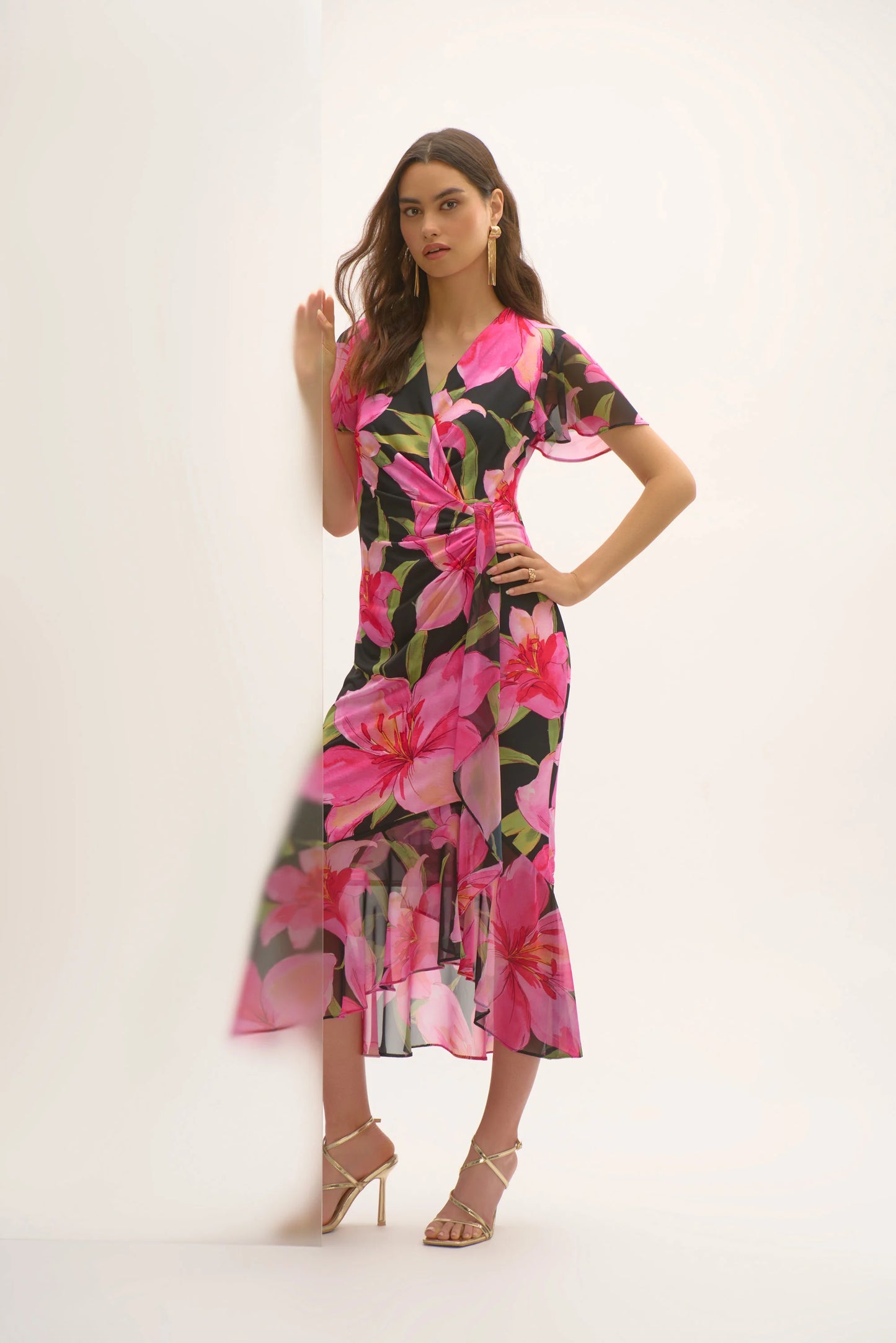 Against a light background, a woman poses in the Joseph Ribkoff Silky Knit and Chiffon Floral Print Wrap Dress, featuring vibrant pink flowers and ruffled chiffon sleeves in a flounce silhouette. She completes the look with strappy heels, one hand on the wall and the other on her hip.