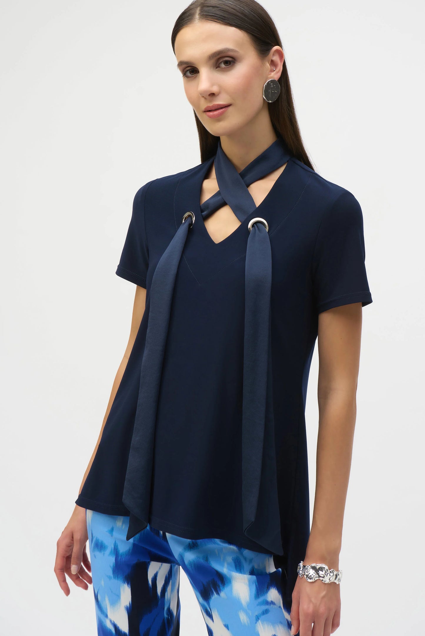 A woman with long brown hair is wearing Joseph Ribkoff's Silky Knit Straight Top with a satin scarf, featuring a unique V-shaped neckline. Her patterned blue and white pants, silver earrings, and bracelet complete the look against a plain white background.