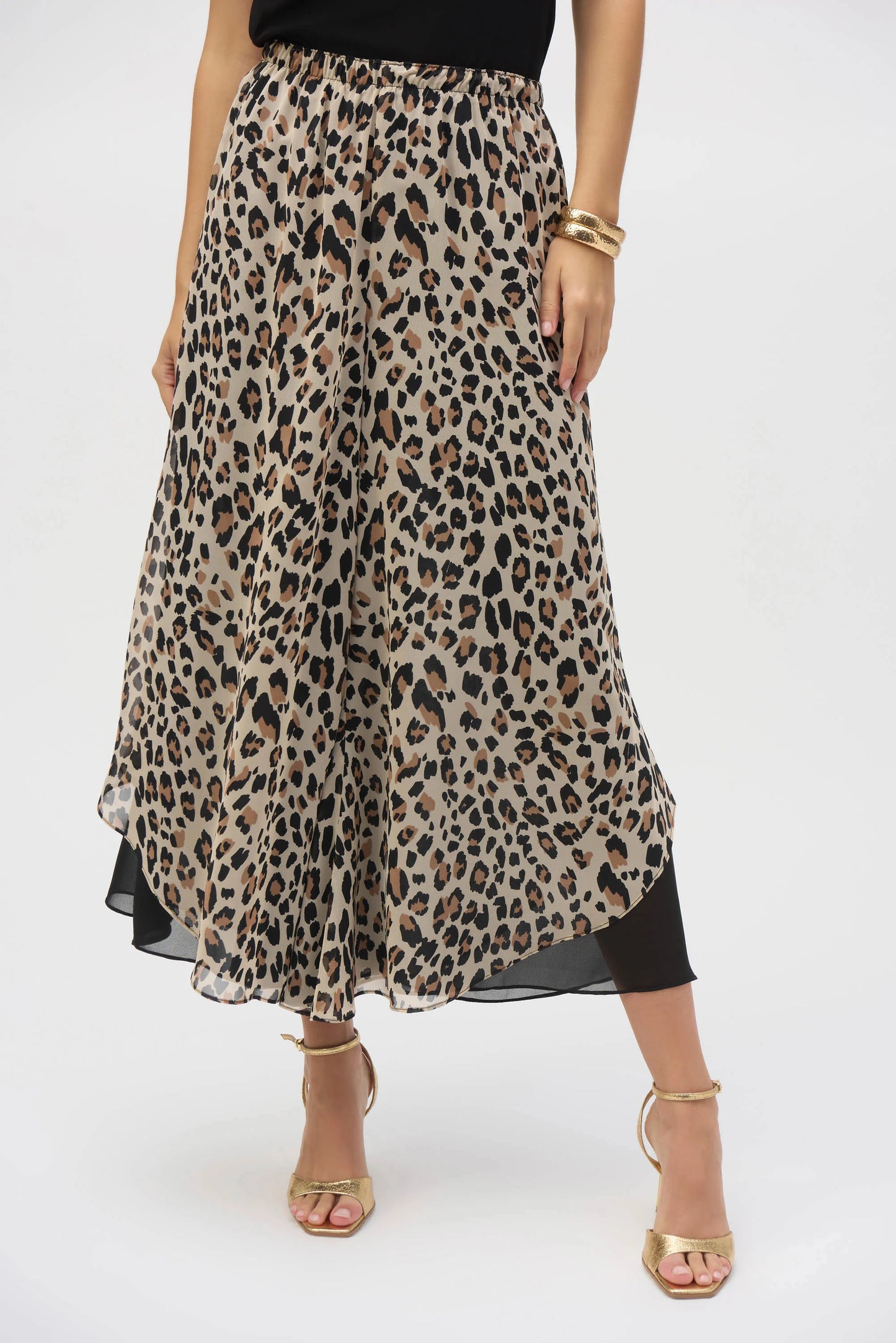 A person stands confidently in Joseph Ribkoff's Chiffon Animal Print Reversible Culotte Pants, paired with a black top, gold bracelets, and chic gold high-heeled sandals. Their hand rests stylishly on their waist against a plain white background.
